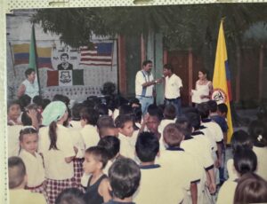 Imagen de un evento escolar con niños reunidos, adultos al frente (uno con micrófono) y una bandera, todo con un fuerte tinte rojizo y la palabra "Izada" en la parte inferior.