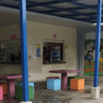 “Zona de cafetería escolar con mesas y asientos de colores bajo un techo cubierto sostenido por columnas azules. Al fondo se observan dos ventanillas de atención de la cafetería y un mural grande con una escena histórica de soldados y una mujer a caballo. A la izquierda, una persona está trapeando el piso.”