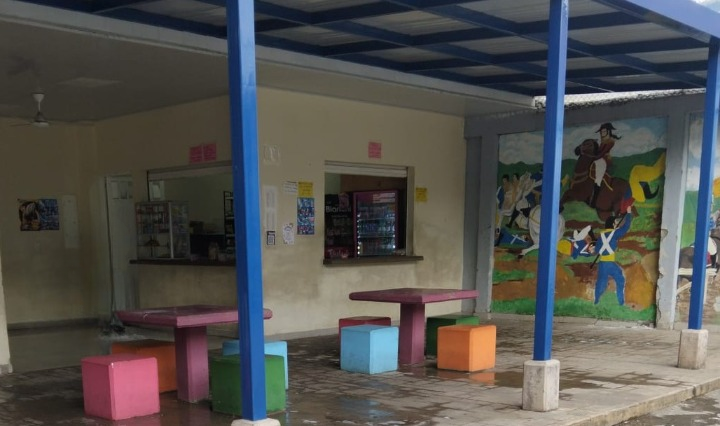 “Zona de cafetería escolar con mesas y asientos de colores bajo un techo cubierto sostenido por columnas azules. Al fondo se observan dos ventanillas de atención de la cafetería y un mural grande con una escena histórica de soldados y una mujer a caballo. A la izquierda, una persona está trapeando el piso.”
