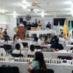 Salón principal de la Institución Educativa John F. Kennedy durante el torneo IRT de ajedrez: decenas de estudiantes y participantes, sentados frente a tableros de ajedrez en largas mesas, mientras al fondo se observa la mesa principal con trofeos, el atril institucional, organizadores y árbitros, y las banderas de Colombia y Boyacá, en un ambiente de competencia académica y formativa.