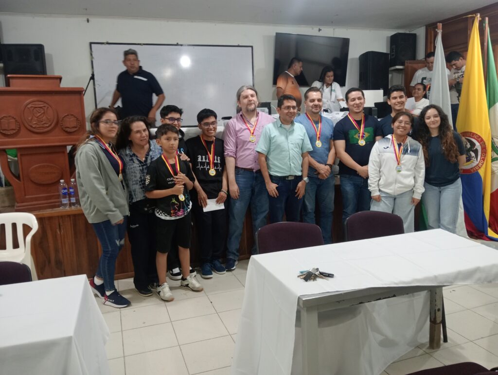 Auditorio de la Institución Educativa John F. Kennedy durante la premiación del torneo IRT de ajedrez: integrantes de la Selección Nacional de Ajedrez de Guatemala, junto a organizadores y participantes locales, posan en grupo portando medallas, con la mesa principal, el atril institucional y las banderas de Colombia y Boyacá al fondo, en un ambiente de reconocimiento deportivo e integración internacional.