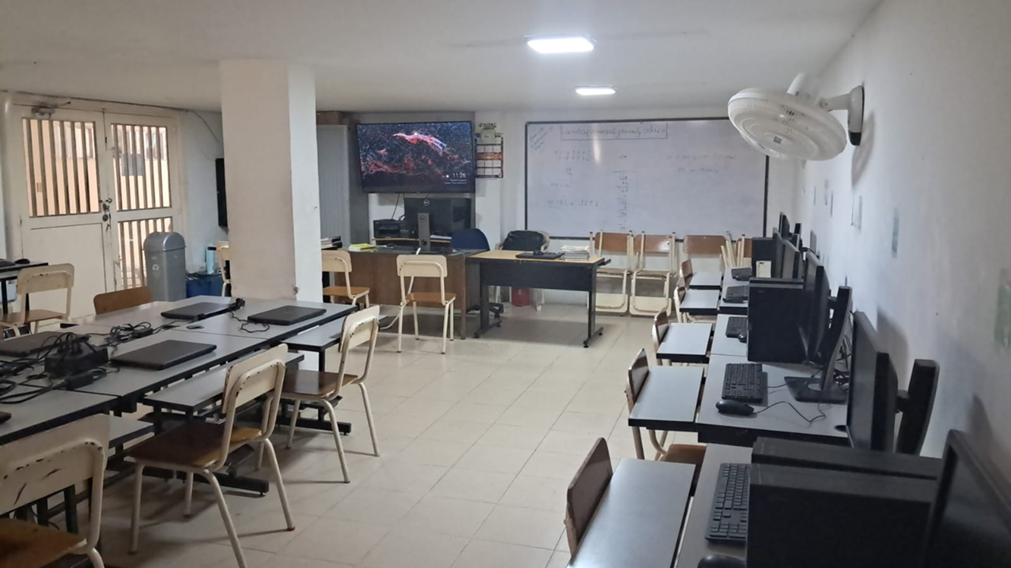 Sala 115 – Informática: aula de informática con computadores de escritorio dispuestos sobre mesas en filas, sillas para estudiantes, cableado organizado, tablero al frente y condiciones adecuadas de iluminación, utilizada para el desarrollo de clases de tecnología e informática en educación básica y media.